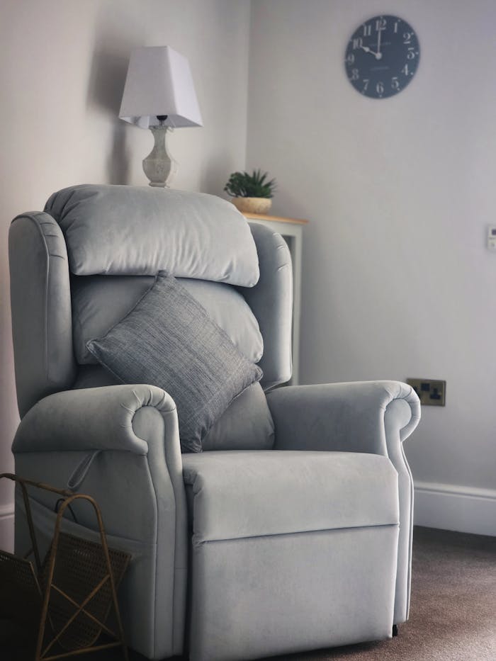 A plush grey armchair with pillow in a serene living room corner, featuring a clock and lamp.