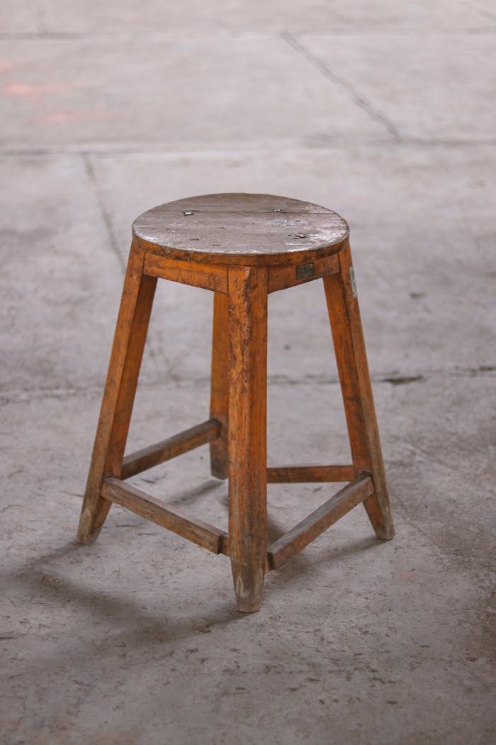 A wooden stool with a rustic finish sits on a concrete floor, displaying a vintage charm.