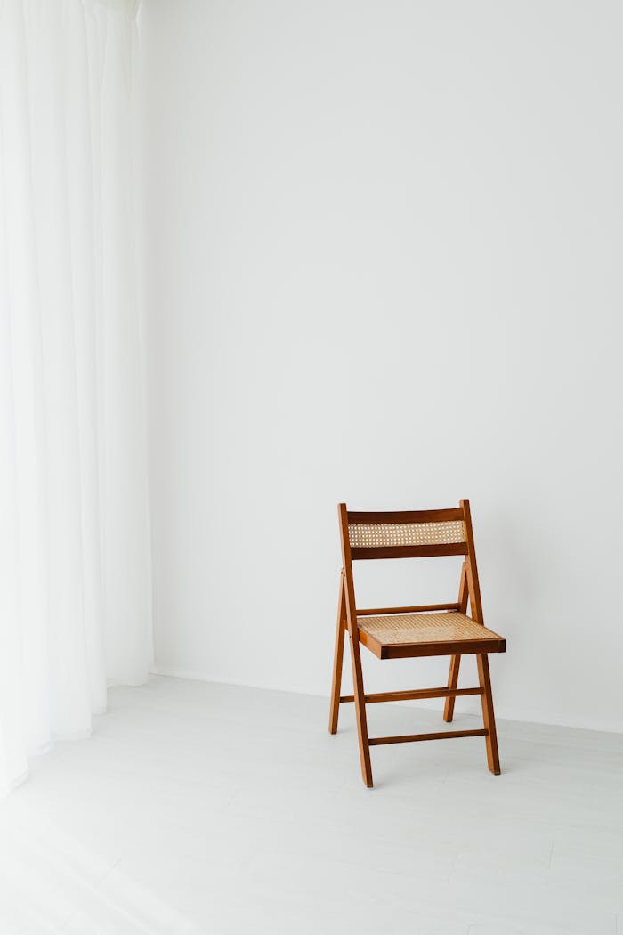 A solitary wooden chair in a minimalist white room with flowing curtains.